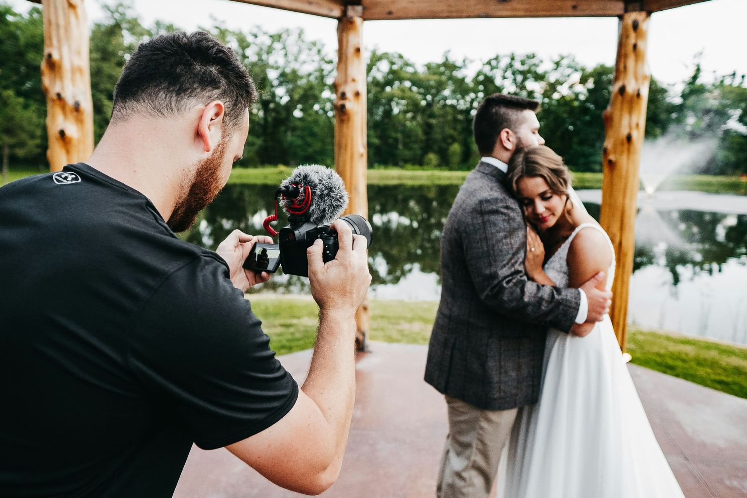 Wedding photographer captures couple embracing under wooden gazebo by serene lake during outdoor ceremony.