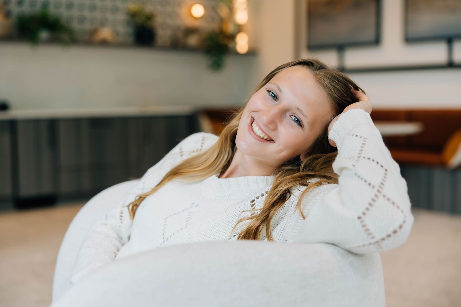 Smiling teenage girl in white sweater relaxing on a couch with hand in her hair.