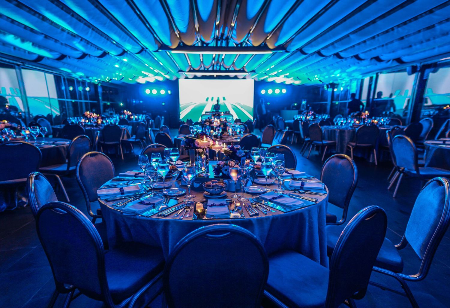Elegant banquet hall with round tables and blue lighting features a large projection screen for corporate events.