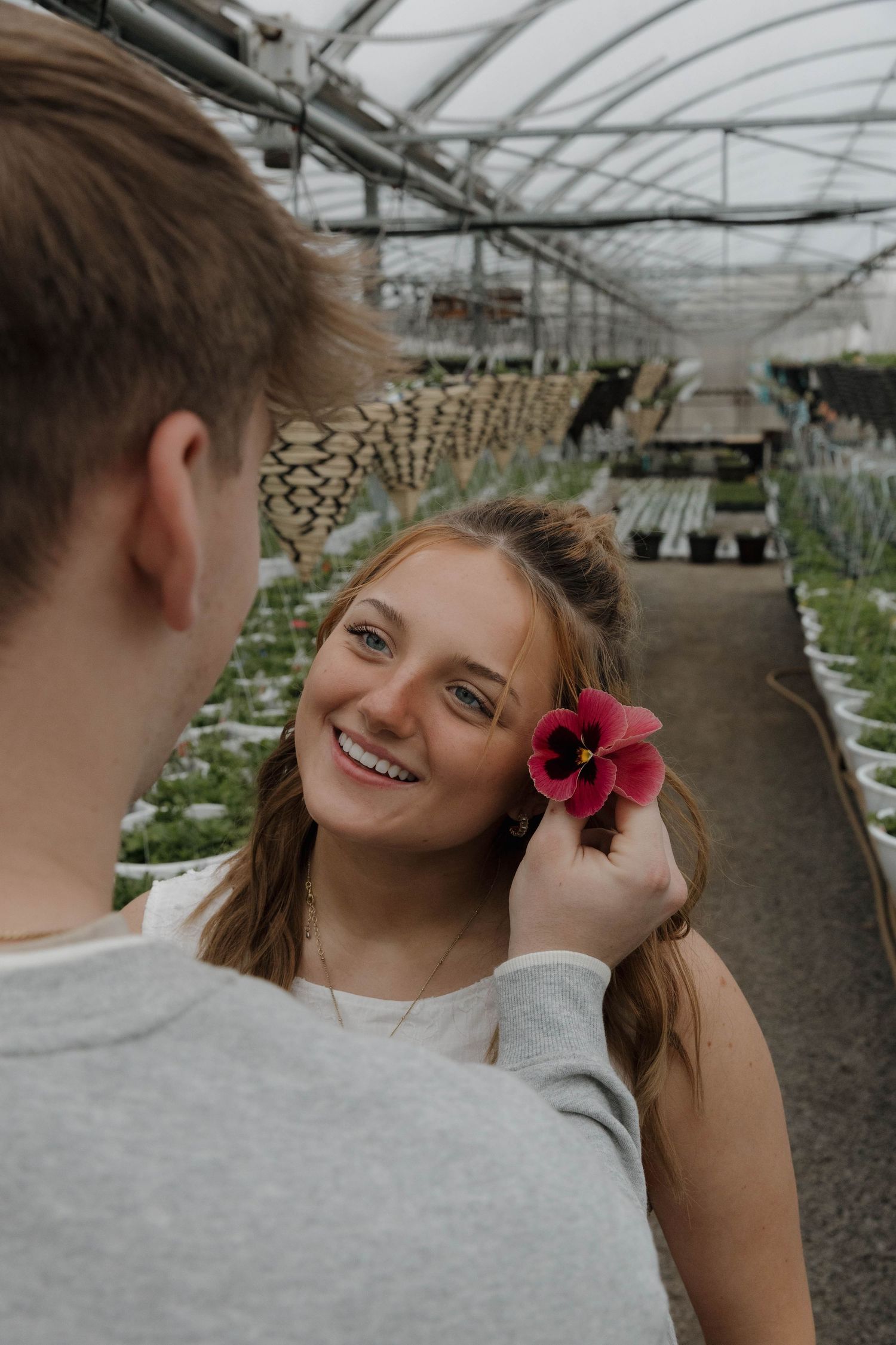 Two people share a sweet moment in a greenhouse as a vibrant pink flower is held up against warm natural lighting.