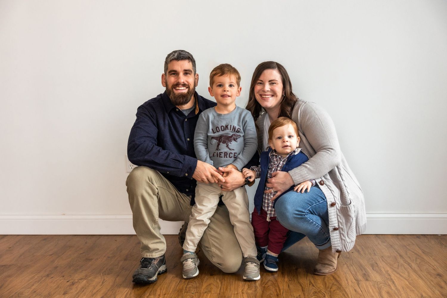 Indoor family portrait against white wall showing warm embraces and casual clothing.