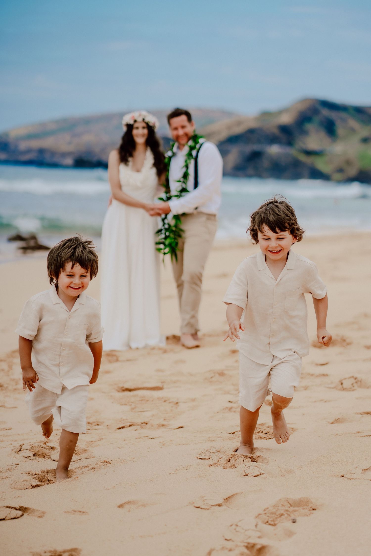 Children playing on a sandy beach while a couple celebrates their wedding with ocean and mountains in background.
