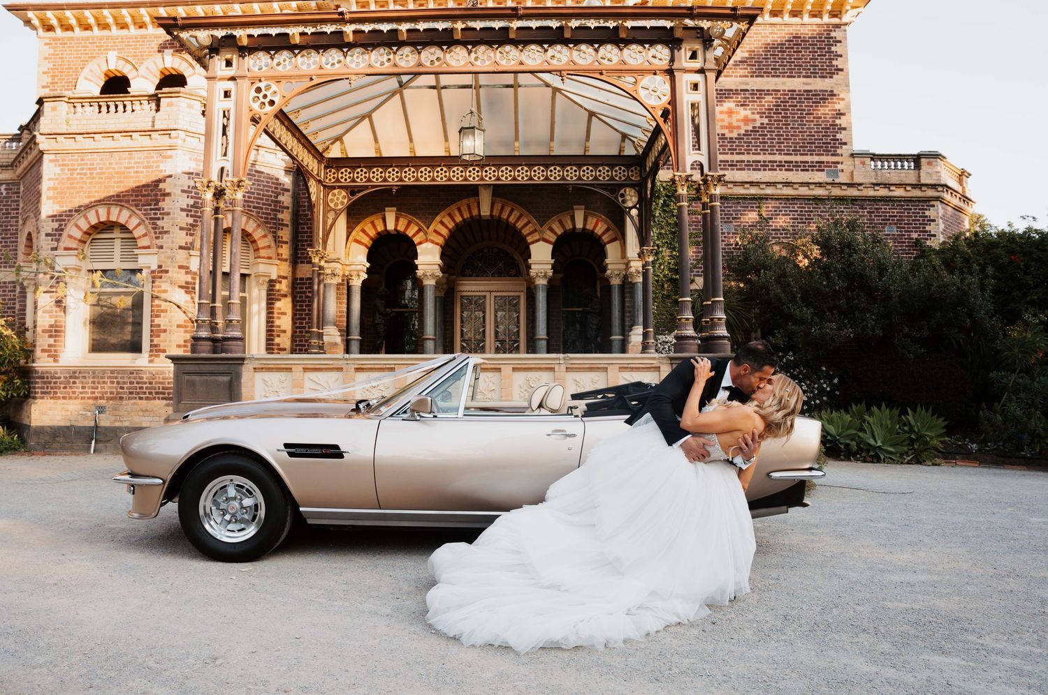 A vintage silver car sits outside an ornate historic stone building with arches at golden hour.