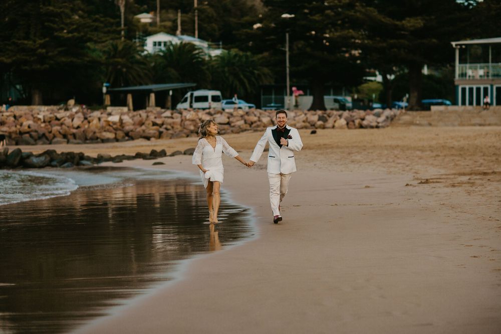 A couple wearing white outfits walks hand in hand along a serene beach at sunset with waves gently lapping at their feet.