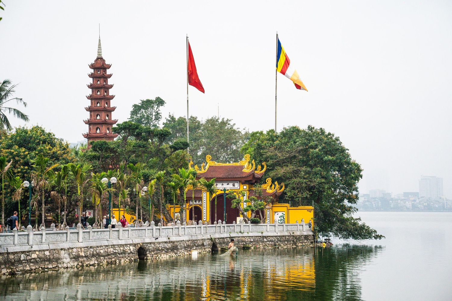 Tran Quoc Pagoda with its iconic red tower reflects in West Lake water with Vietnamese and Buddhist flags in Hanoi, Vietnam.