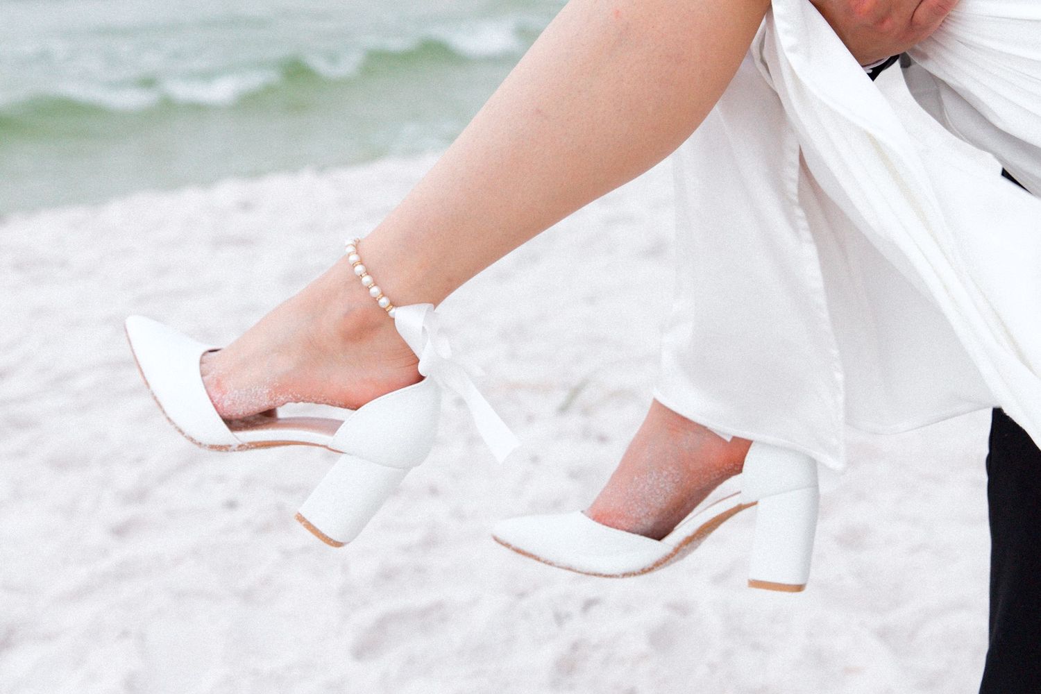 Detail shot of bride's white block heel sandals against beach sand.