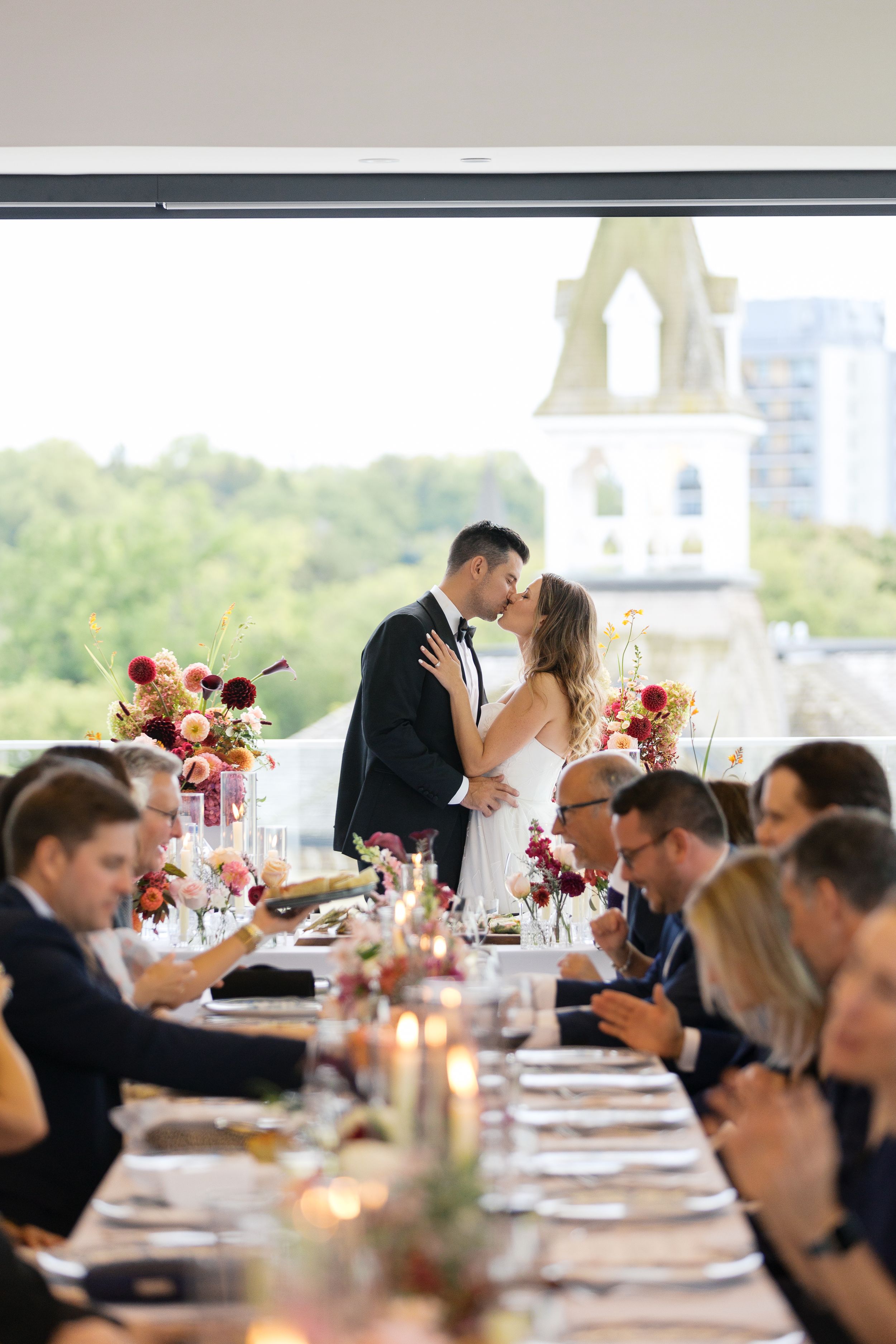 Elegant wedding reception with couple kissing while guests dine at long table.