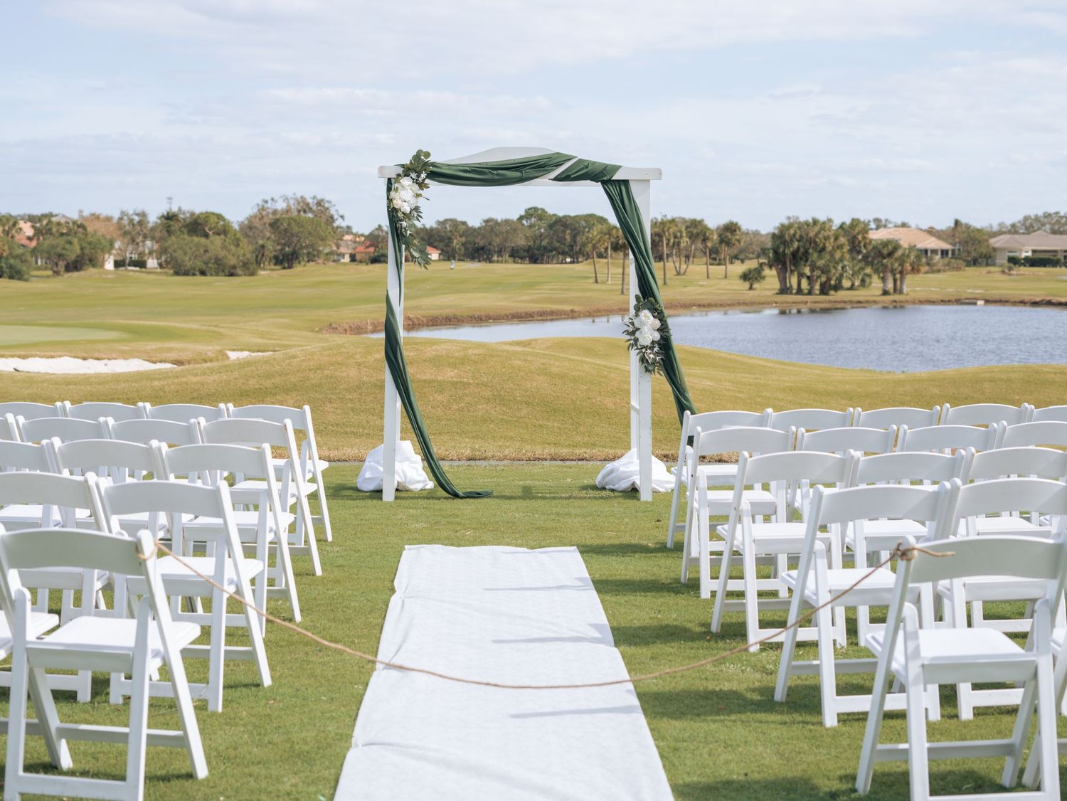 White aisle runner leading to a green-draped arch with white chairs on a golf course.