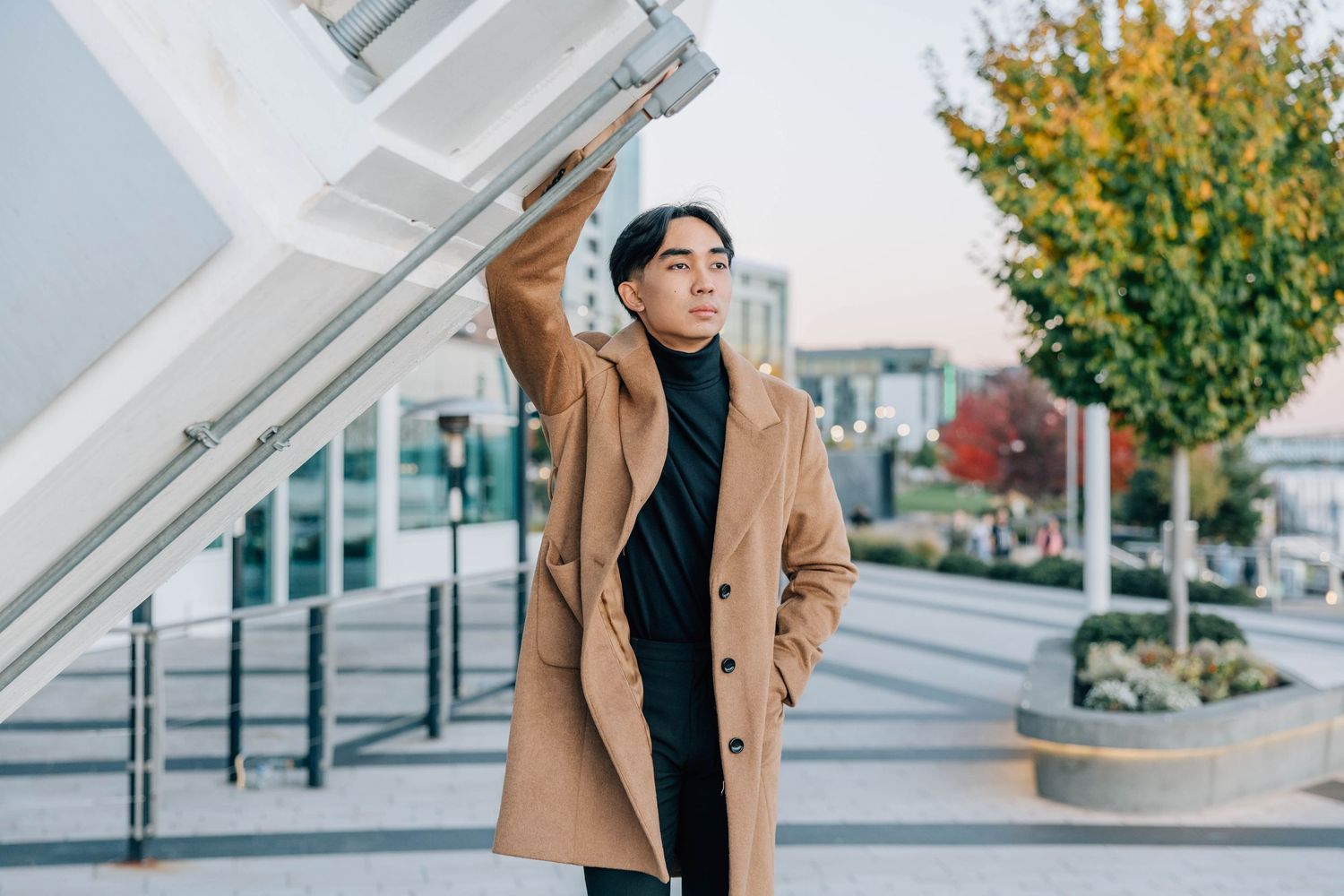 Stylish young man in camel coat posing near modern architecture in urban setting.