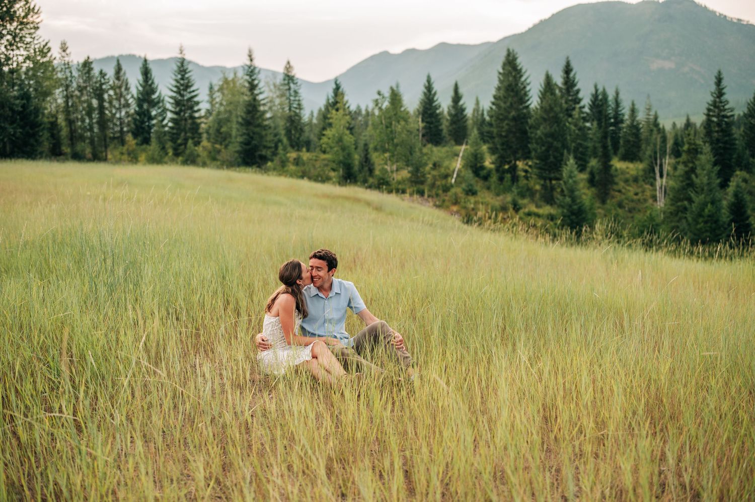 A couple sits together in a scenic mountain meadow surrounded by tall grass and evergreen trees in the distance.