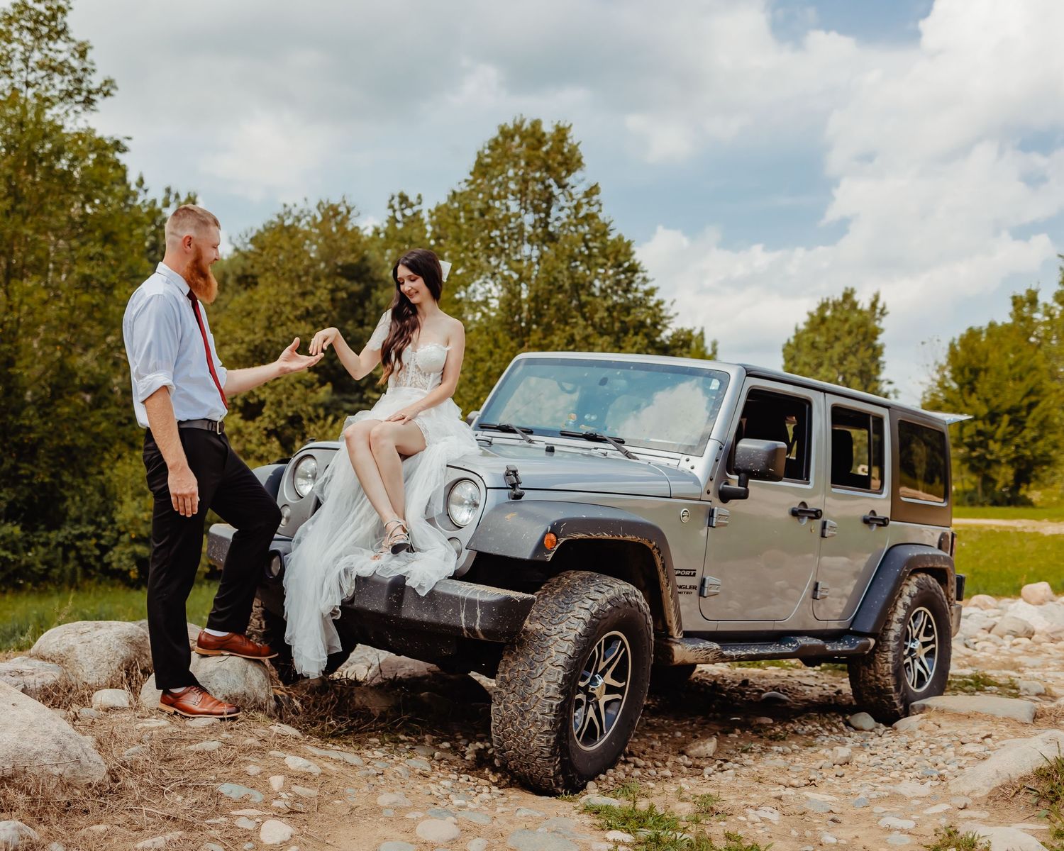 Wedding couple posing with grey Jeep Wrangler on rocky outdoor terrain.