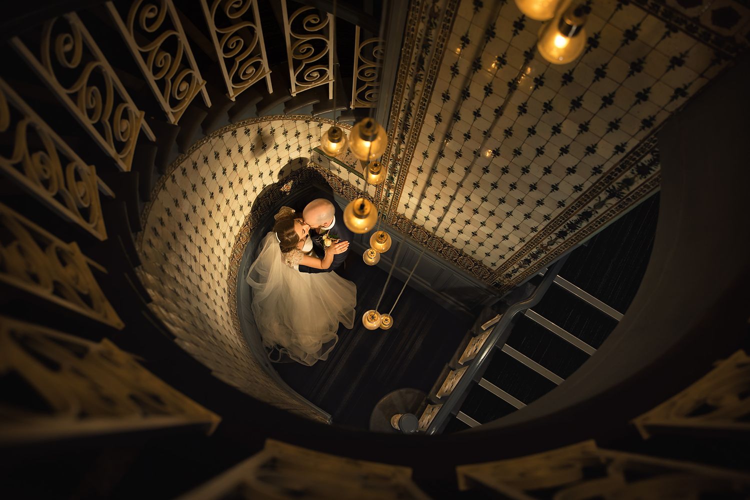 Overhead spiral staircase view of flowing white wedding dress against ornate patterned ceiling and railing.