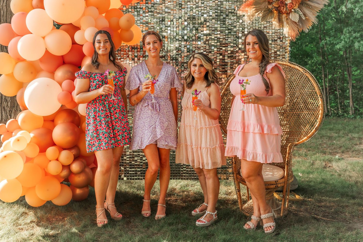 Four people pose against an orange balloon arch with a peacock chair.