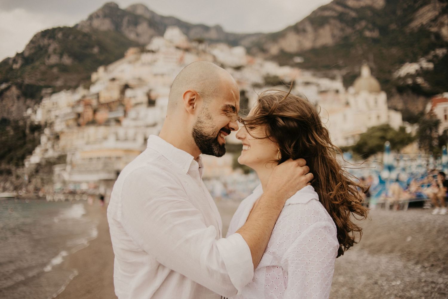 Romantic couple embracing on beach with Positano coastal town behind them.