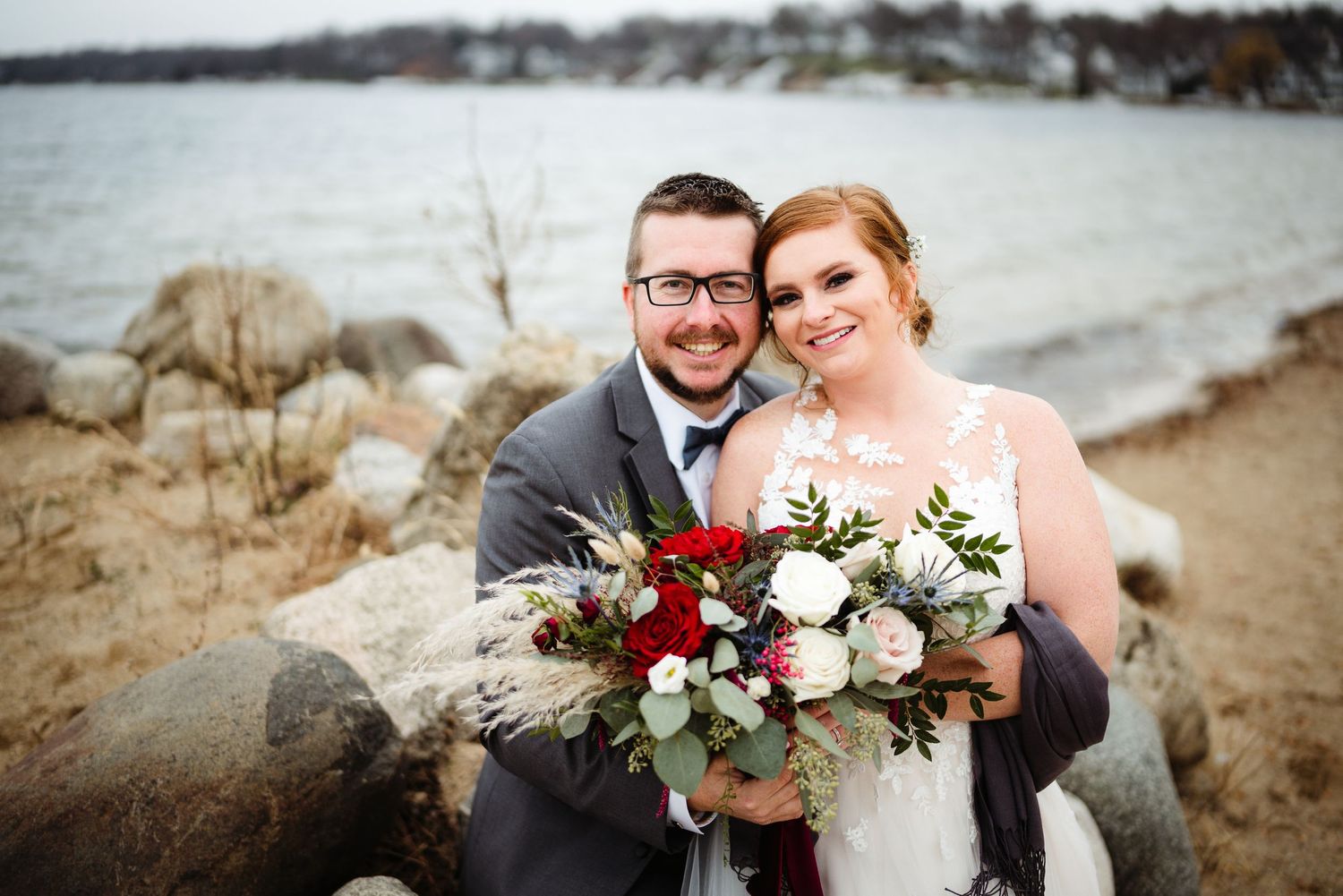 A wedding couple poses near a rocky beach shoreline with a vibrant red and white floral bouquet.