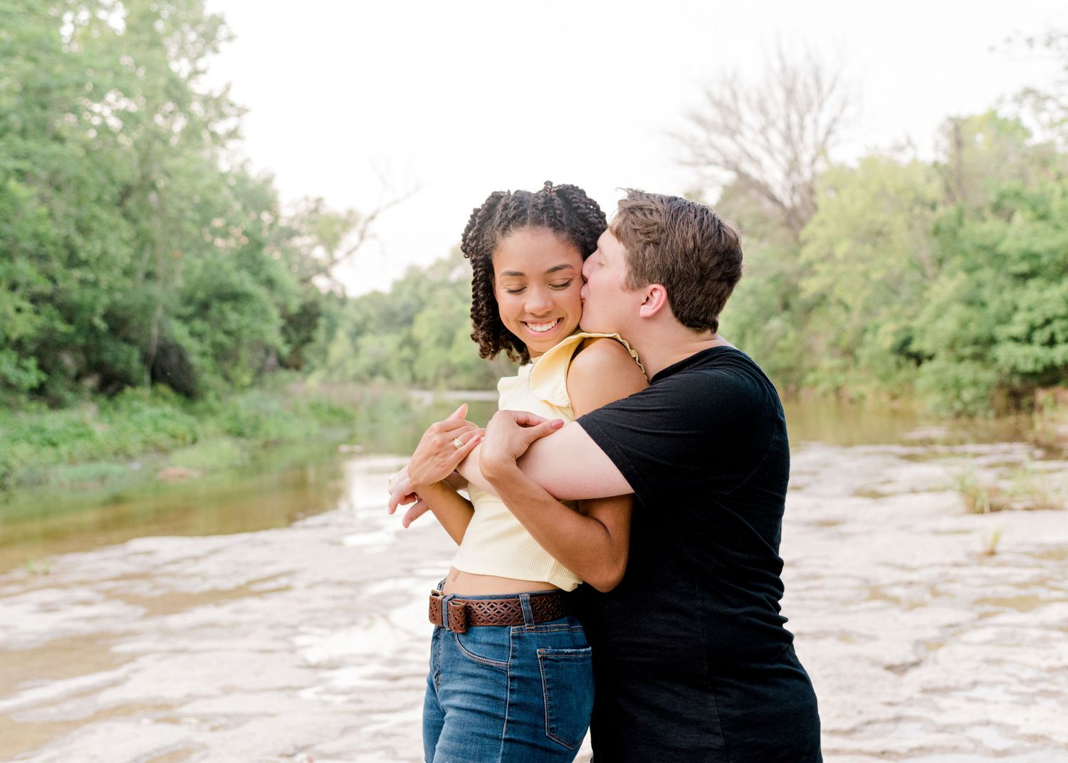 engaged couple in austin, tx. guy's arms are wrapped around girl as he kisses her cheek.