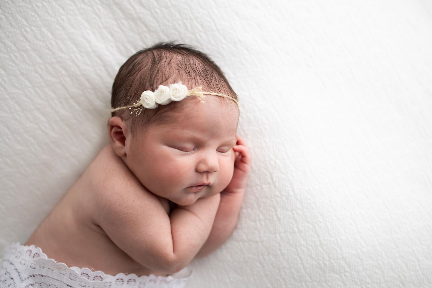 Newborn baby girl laying on white blanket with floral headband.  Both hands under her cheek.