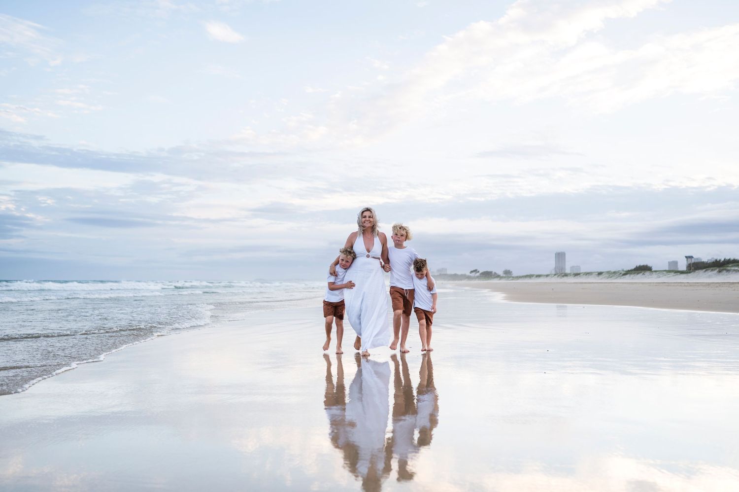Family silhouettes holding hands on reflective beach at sunset with cityscape in background.
