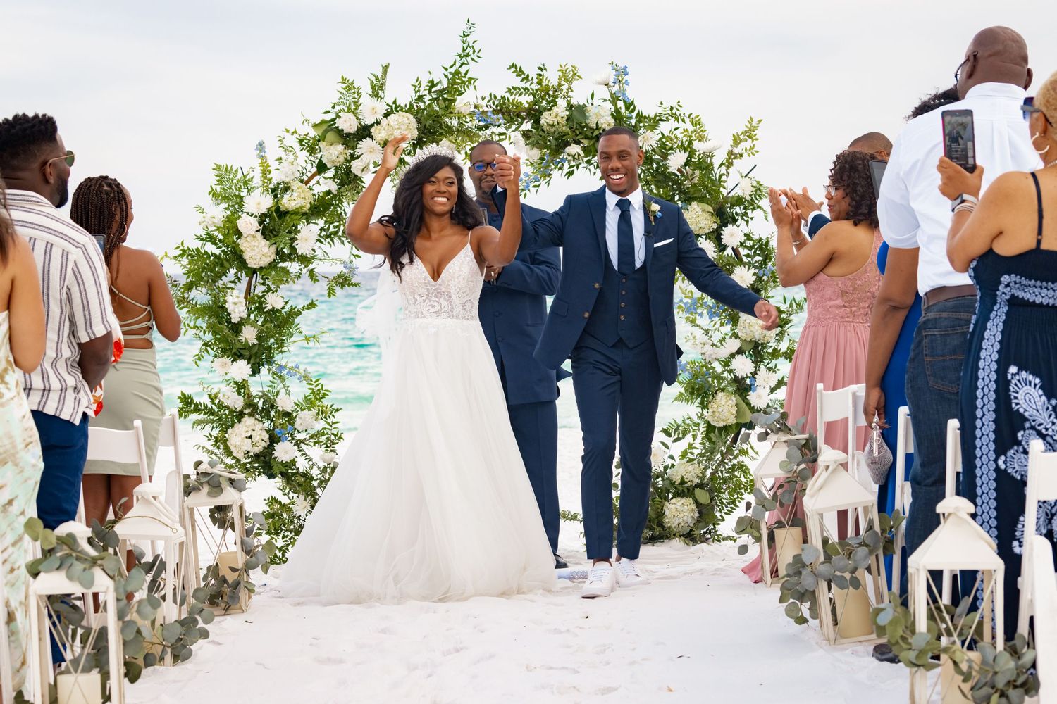 Jubilant newlyweds exit their wedding ceremony under a lush white floral arch while guests cheer.
