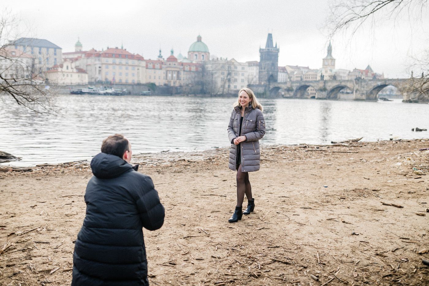 A couple standing on a sandy riverbank with the iconic Charles Bridge and Prague skyline in the background.
