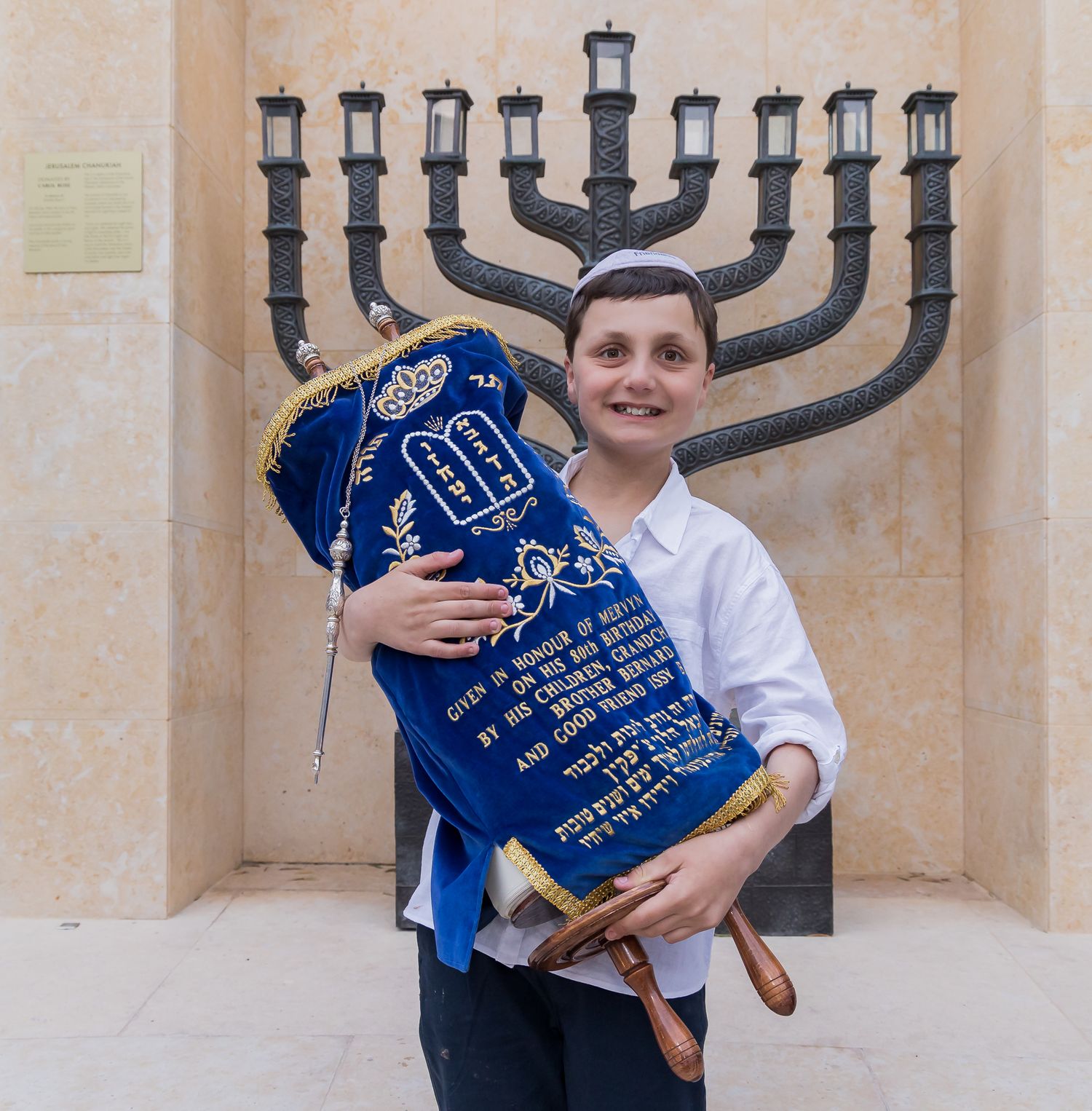 A person holds a decorative blue Torah scroll in front of a large menorah display.