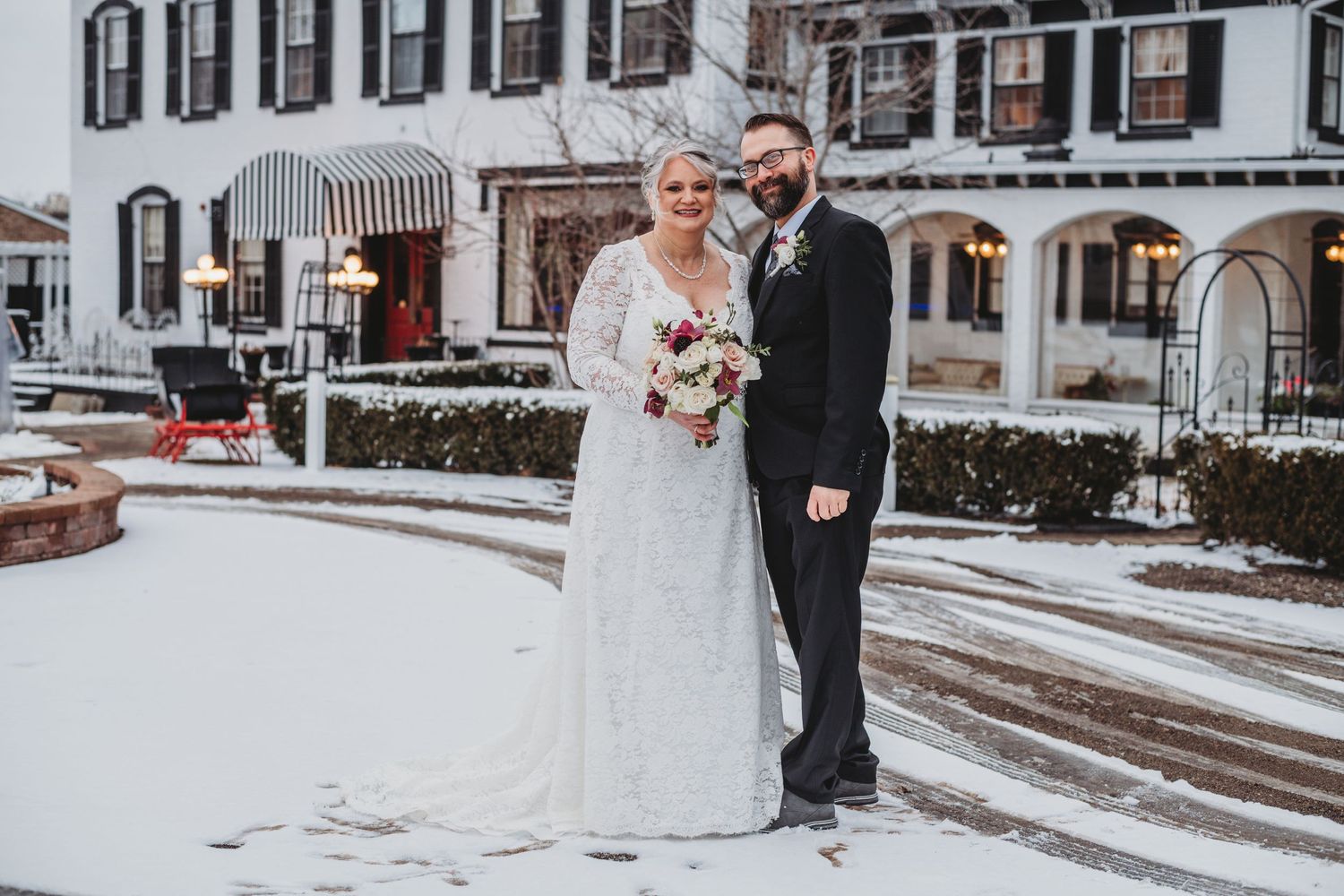 A winter wedding portrait outside a historic white building with snow on the ground.
