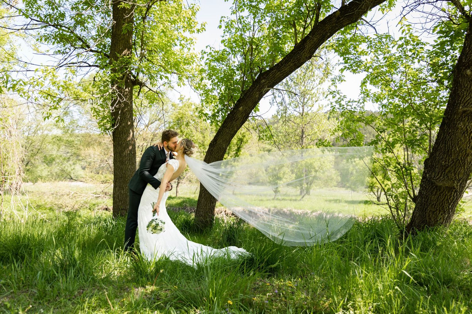 A bride and groom share a romantic kiss under lush green trees, with her veil flowing in the breeze.