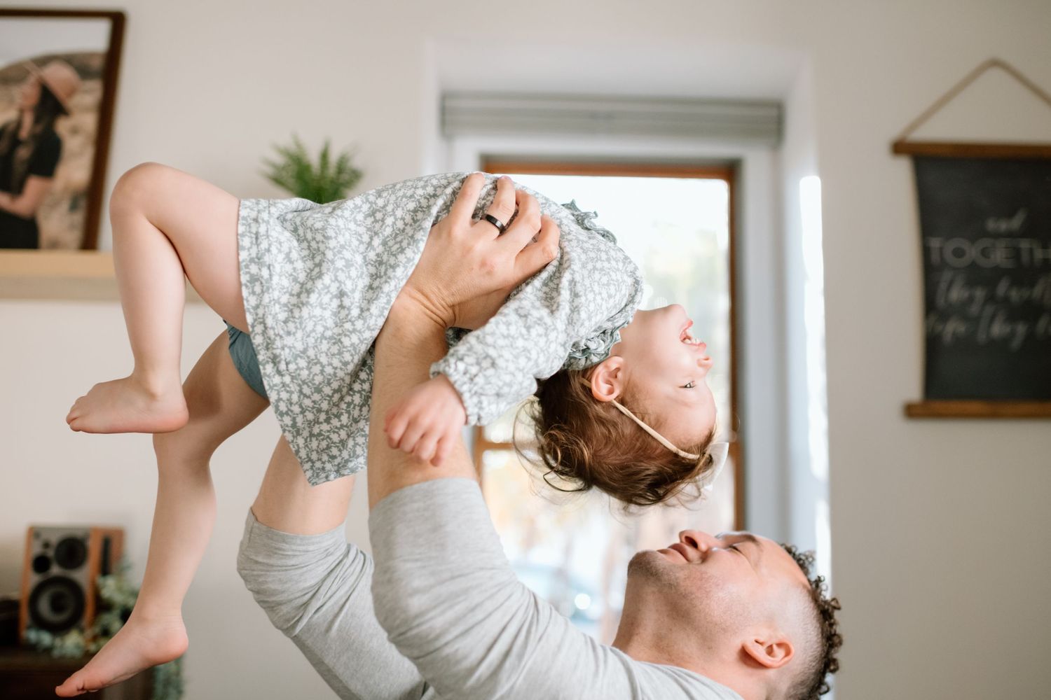Parent playfully lifting child in air against bright window in home.