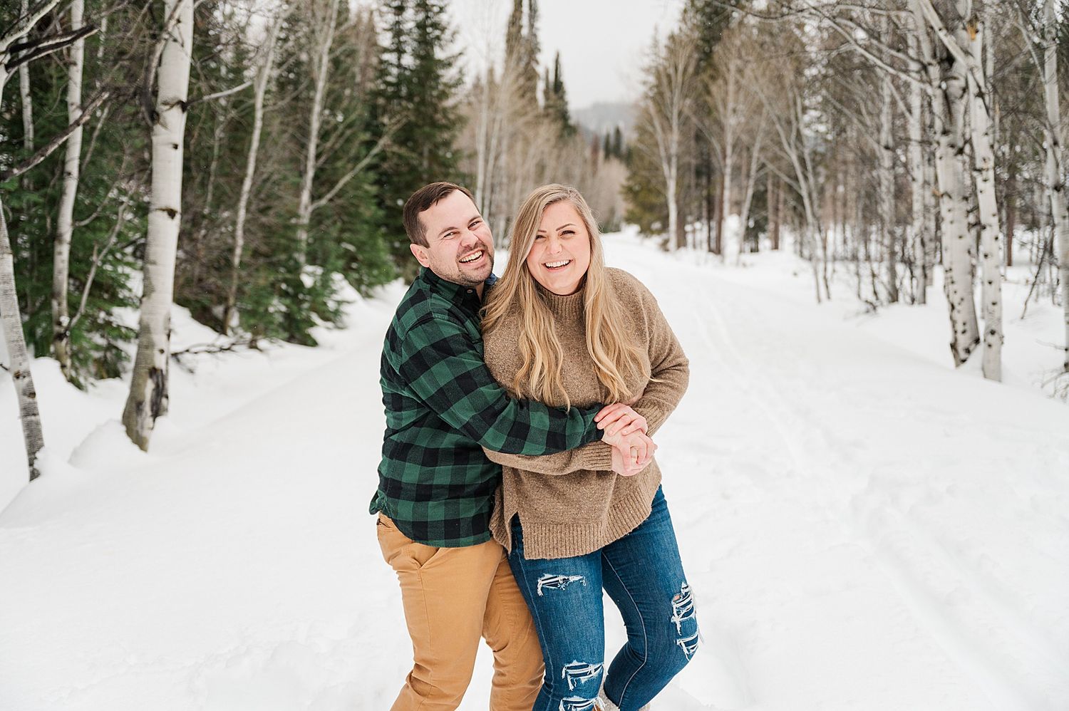 A couple laughs together while embracing on a snow-covered forest path.