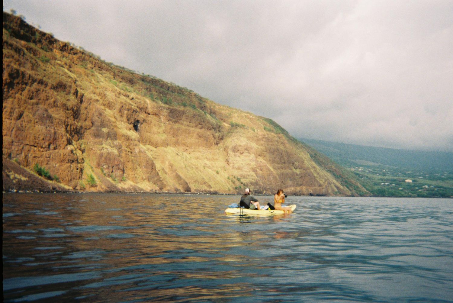 couple sitting on a kayak in the ocean, the woman is taking a picture of something, a mountain rises behind them