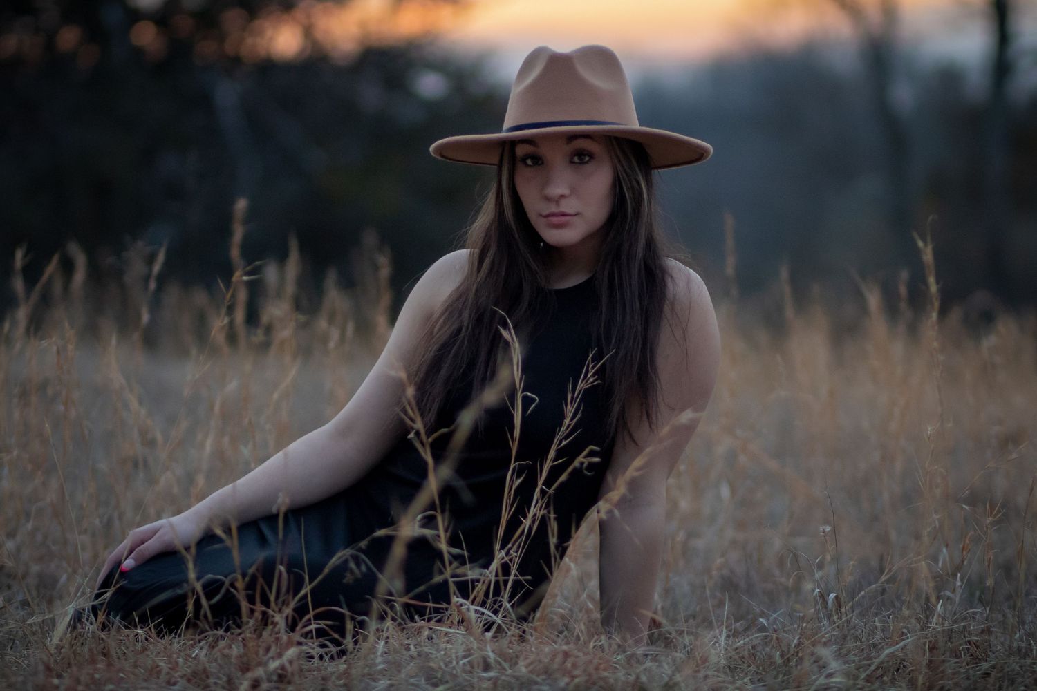 Young woman in a wide-brim tan hat sits in a golden grass field at sunset wearing a black dress.