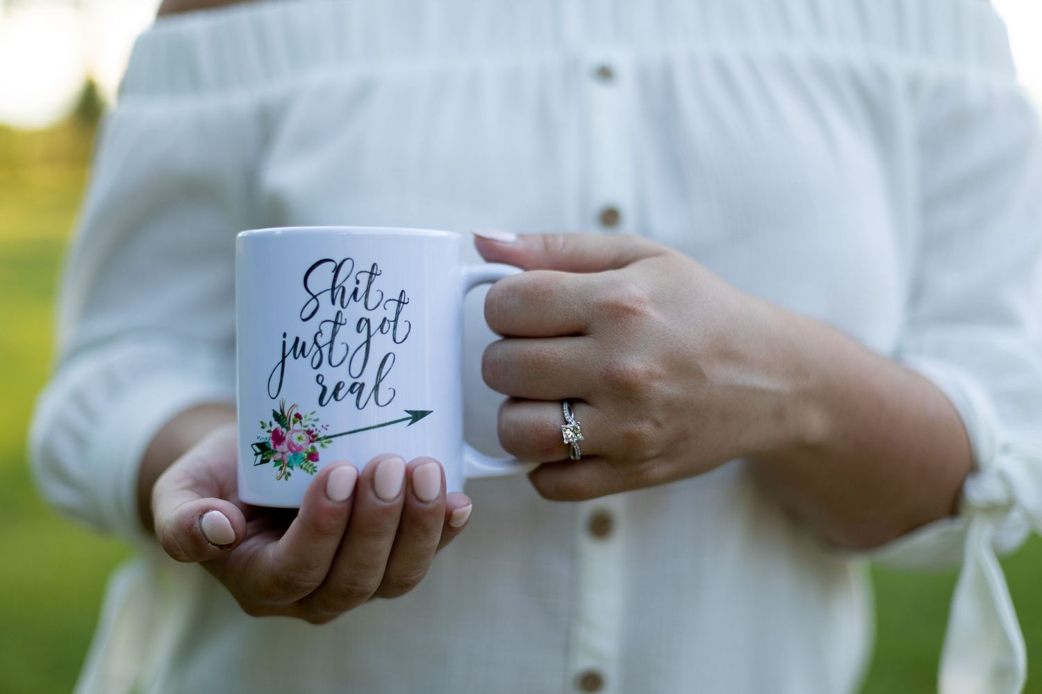 White coffee mug with handwritten text shown in outdoor setting with engagement ring visible.