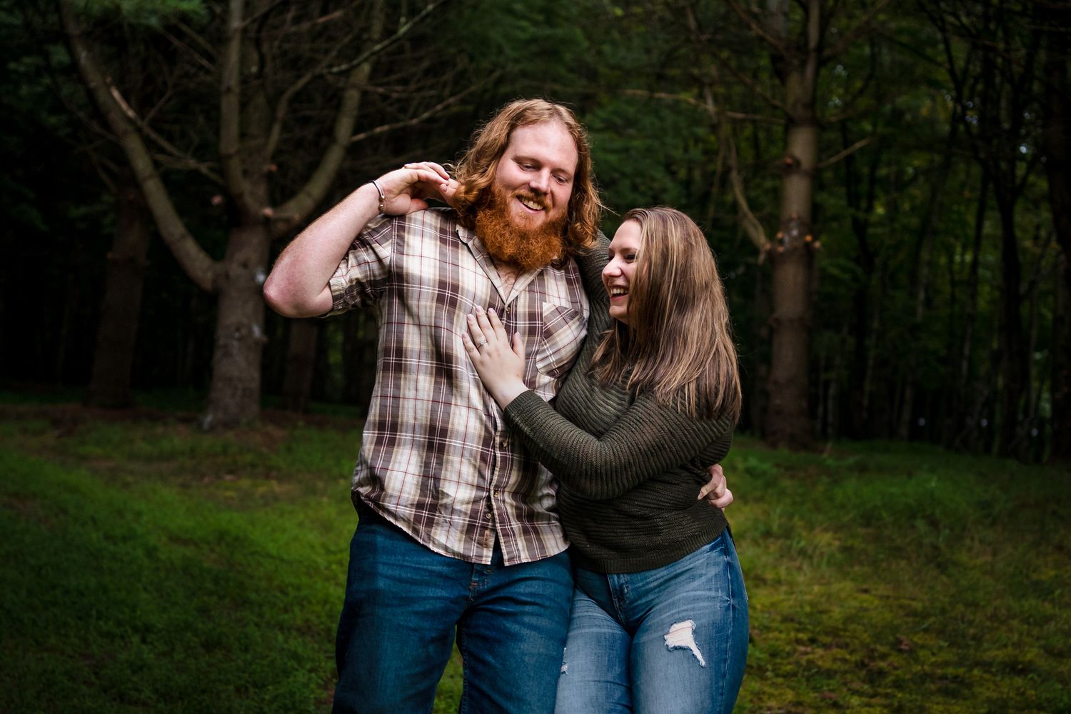 A couple in casual attire embraces while walking through a wooded outdoor setting.