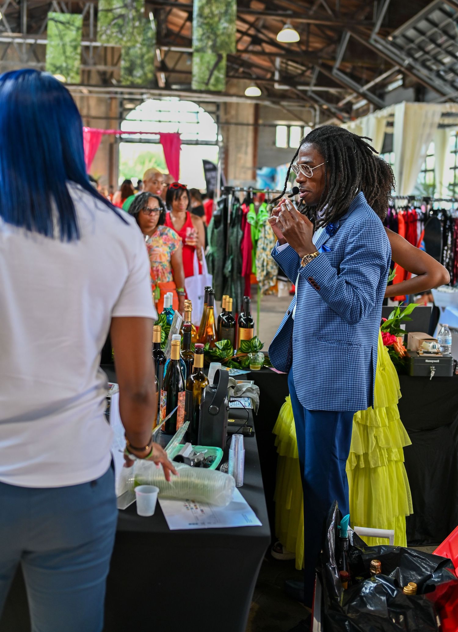 Vendor interacts with customer at indoor market booth with clothing racks and merchandise displays.