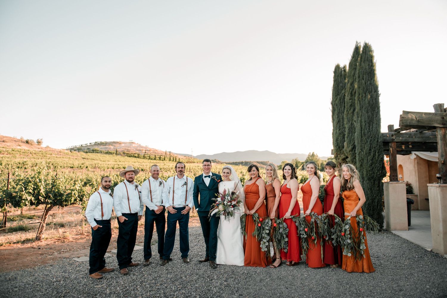 Wedding party poses in formal attire against scenic vineyard backdrop at sunset.