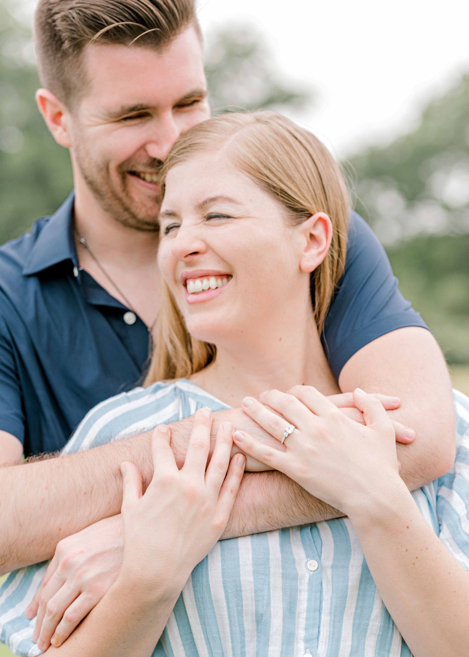 engaged couple smiling together. guy's arms wrapped around girl's shoulders. her new ring on display.