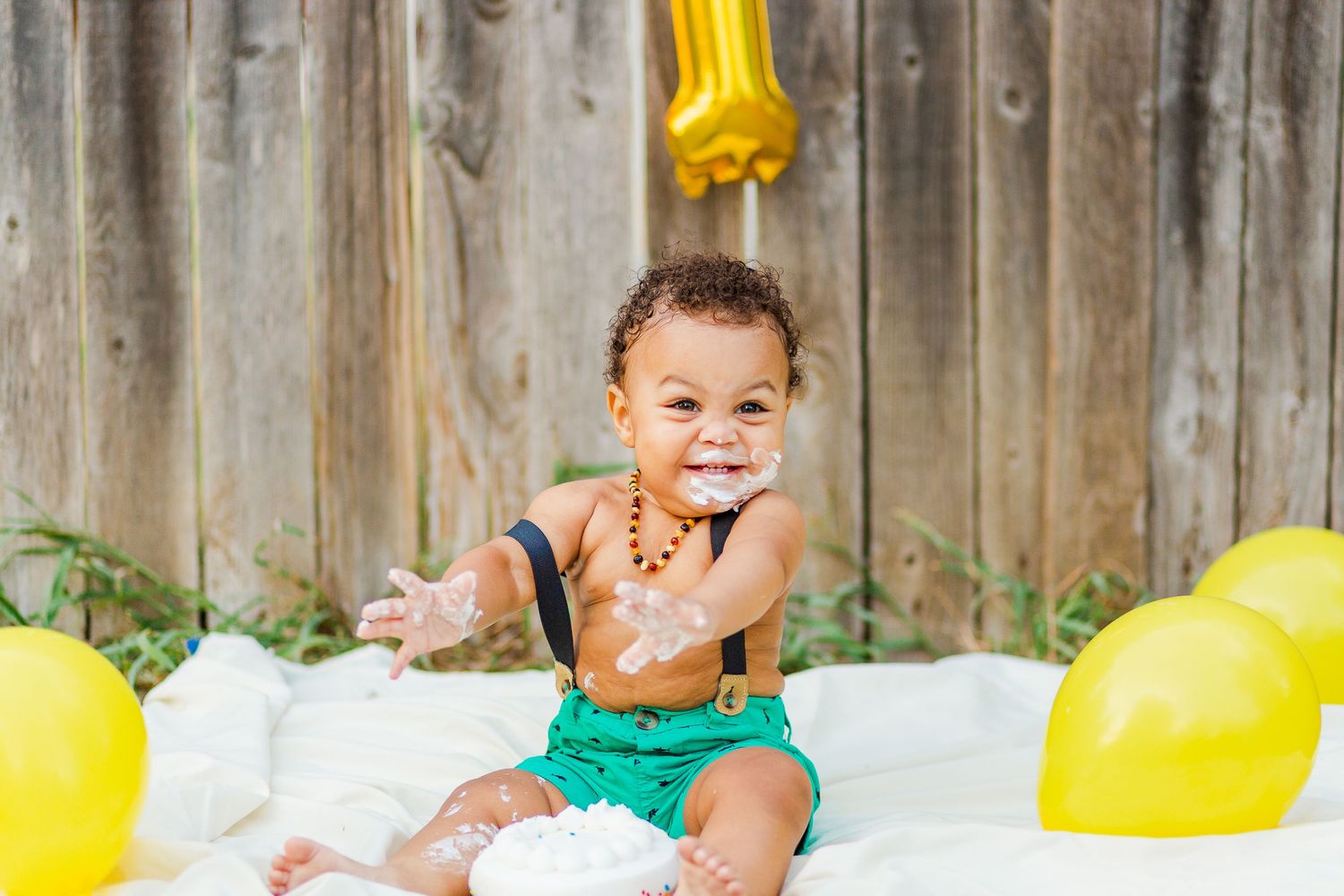 Happy toddler at first birthday shoot with cake and yellow balloons against wooden fence.