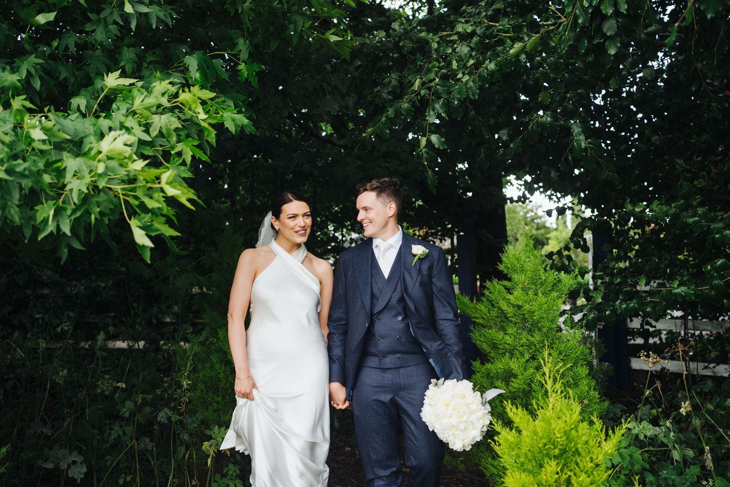 Newlyweds walking together through lush garden greenery in formal wedding attire.