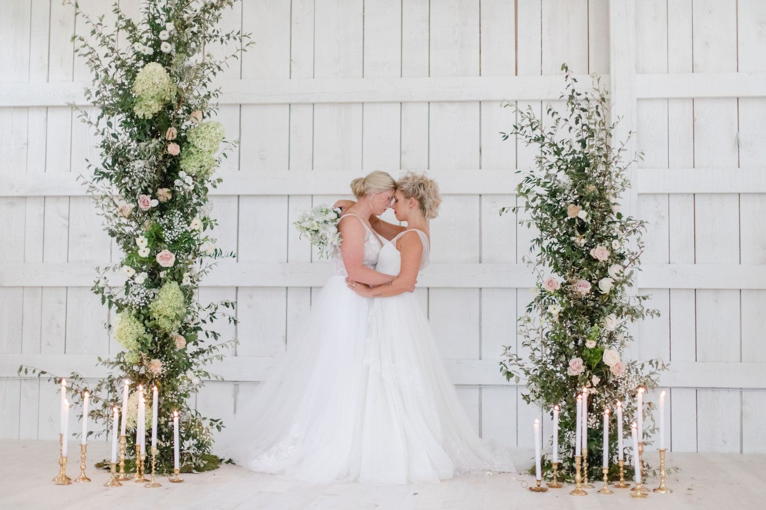 White floral wedding arch with greenery against a rustic white paneled wall.