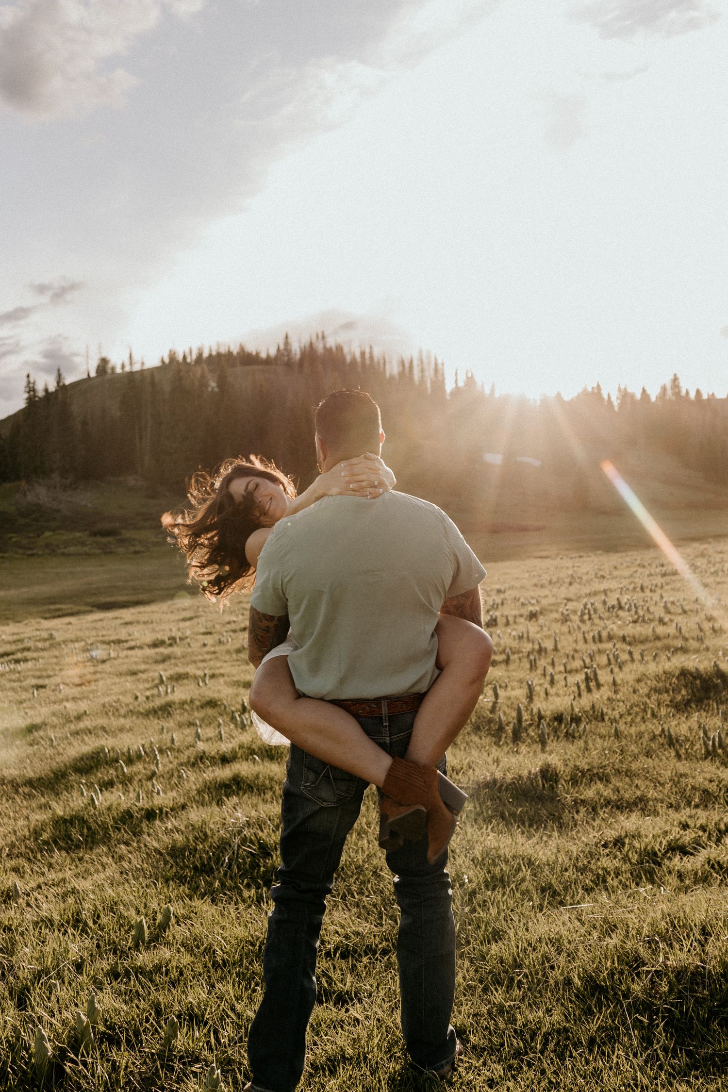Couple embracing in a sunlit mountain meadow at golden hour with forest backdrop.