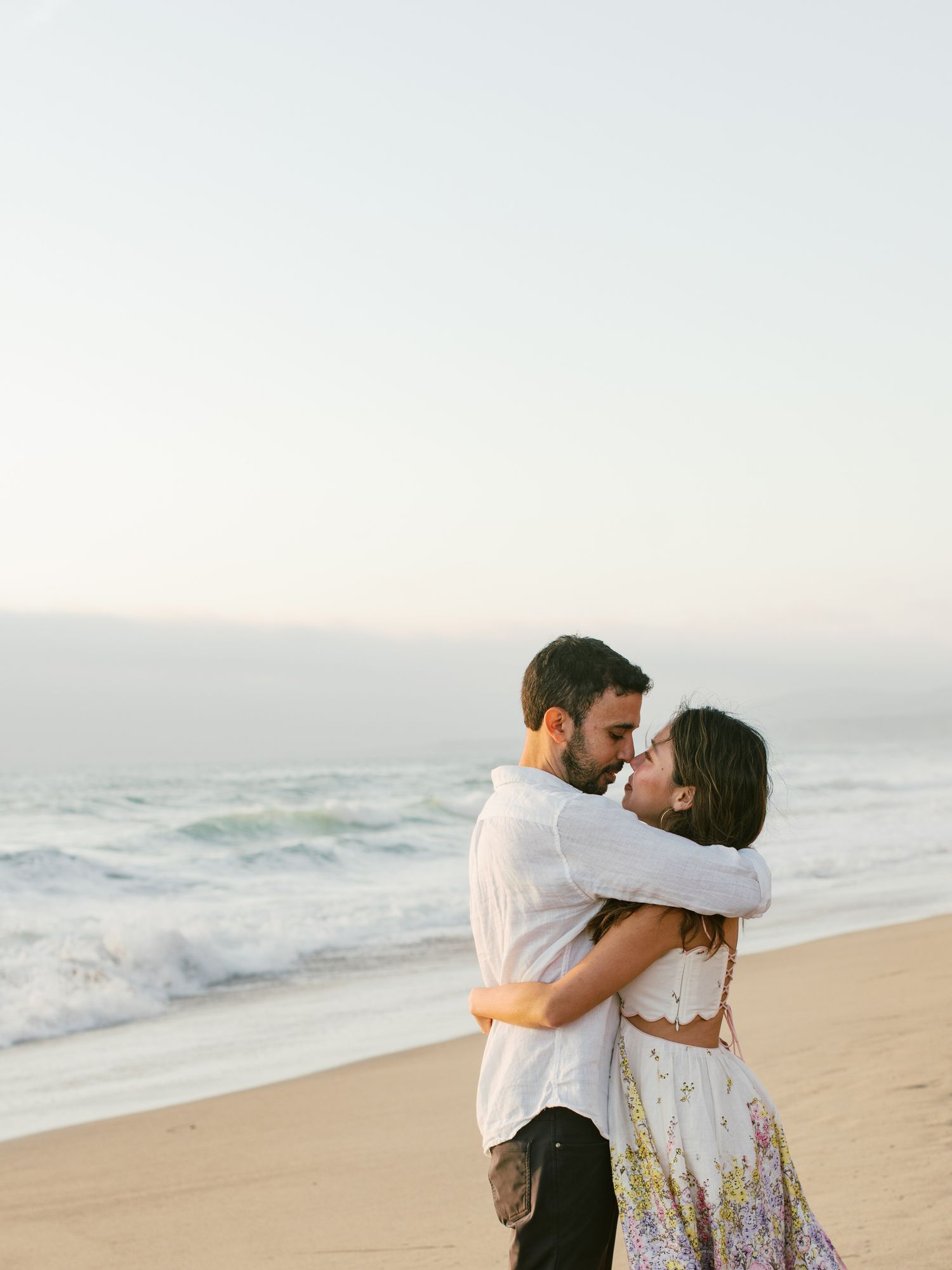 Couple embracing on beach at sunset with ocean waves in background.