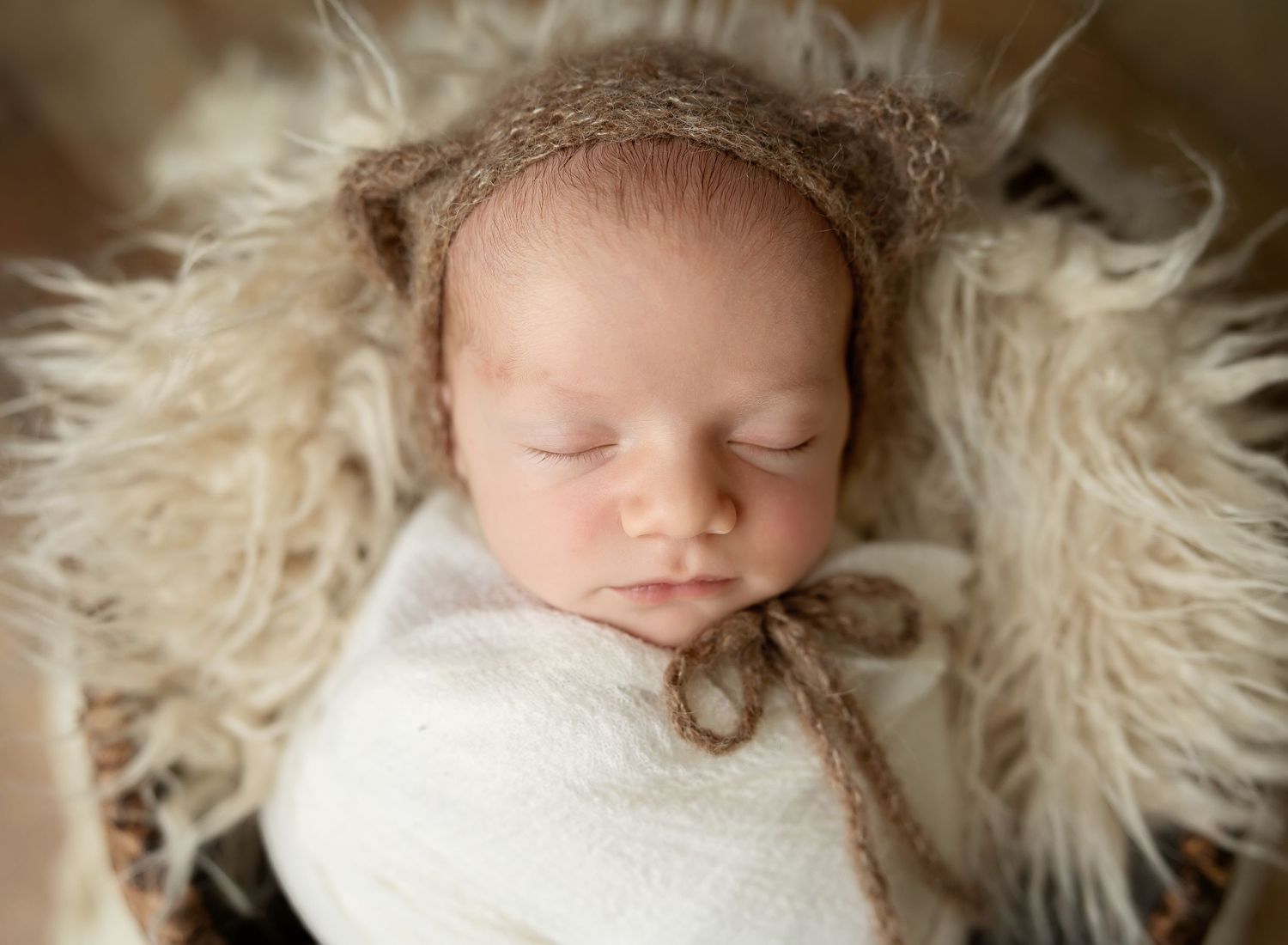 Sleeping newborn wrapped in white swaddle on cream fur backdrop.