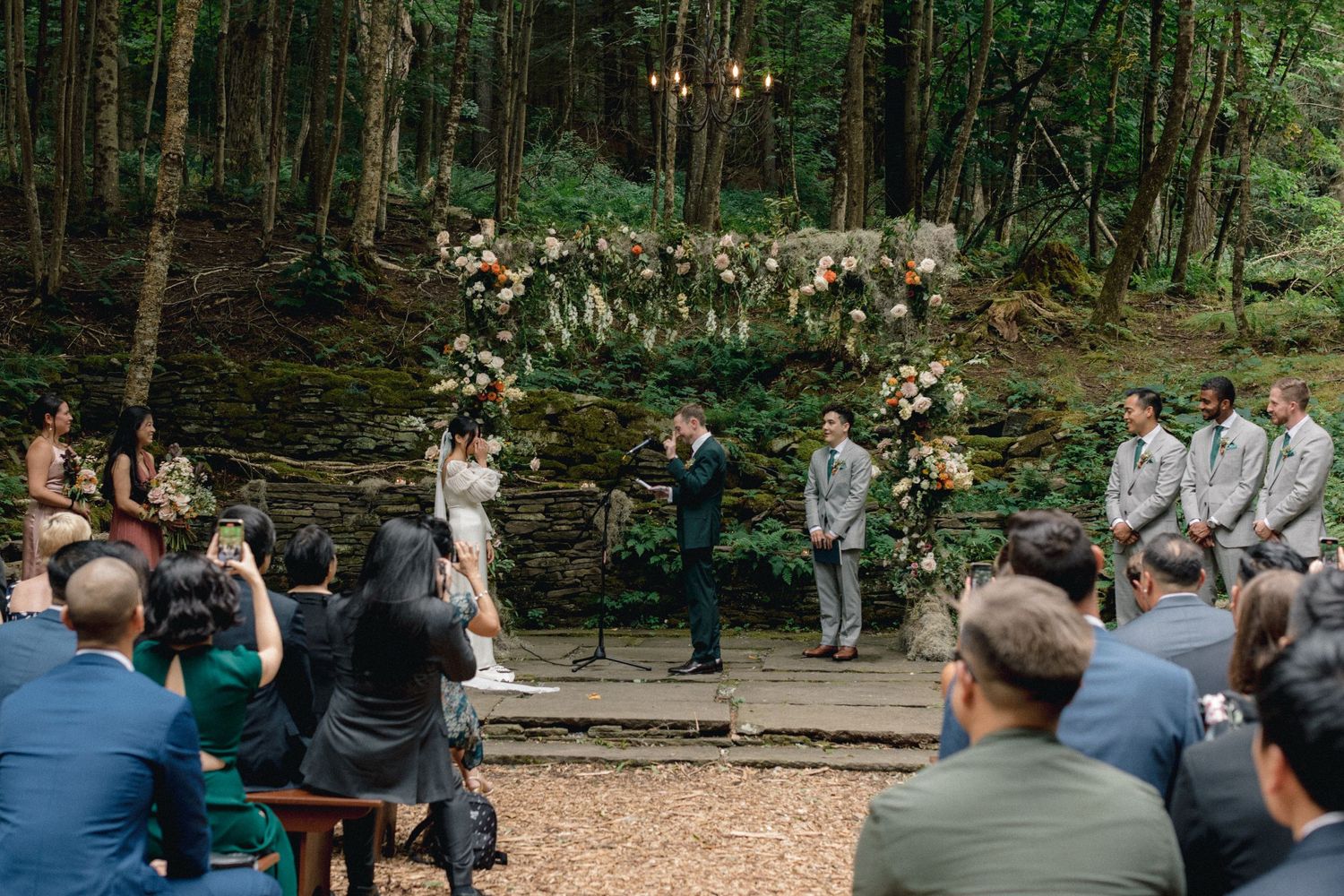 Outdoor wedding ceremony in wooded setting with floral arch and seated guests.