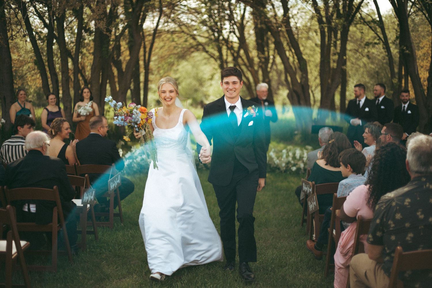A wedding couple walks down the aisle after their outdoor ceremony in a wooded setting.