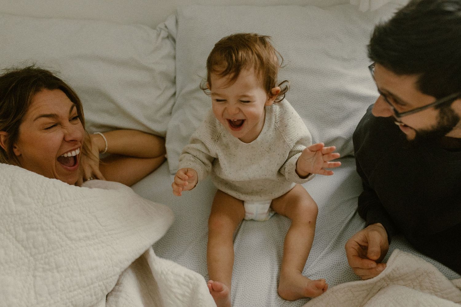 A joyful moment captured on a bed with a small toddler in white being playfully entertained.