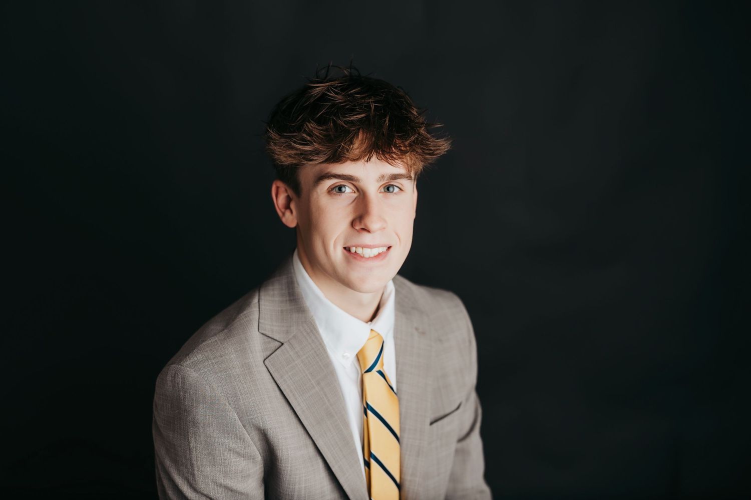 Professional portrait against dark background featuring light gray suit and yellow striped tie.