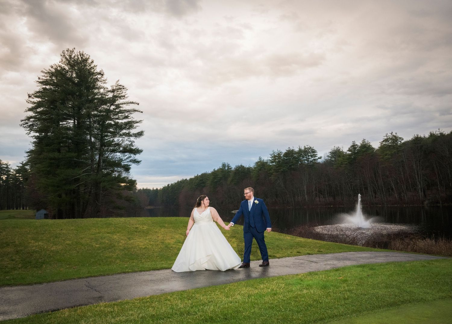 Couple walks along path with pine trees and fountain in background during wedding photoshoot.