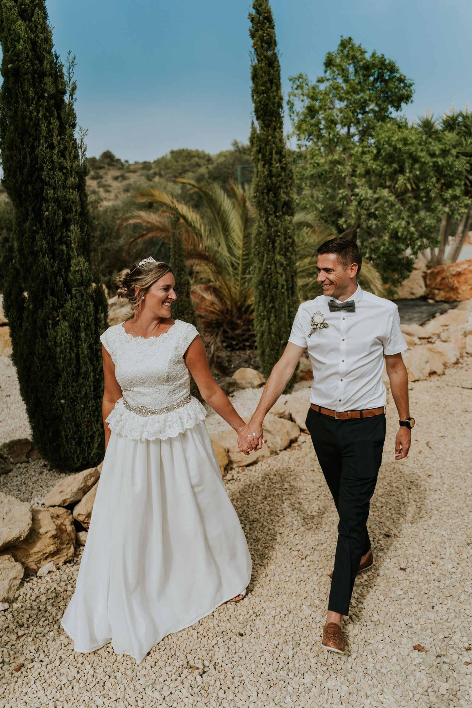 A couple walks hand in hand through a cypress tree-lined path in summery wedding attire.