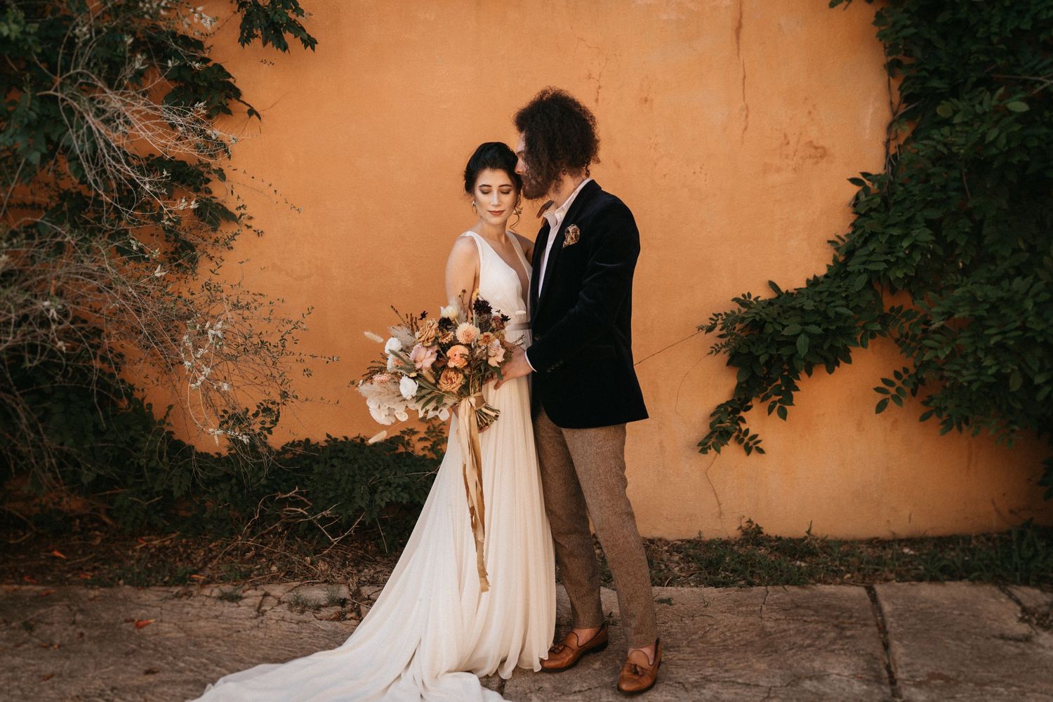 Wedding couple embracing against terracotta wall with lush florals.