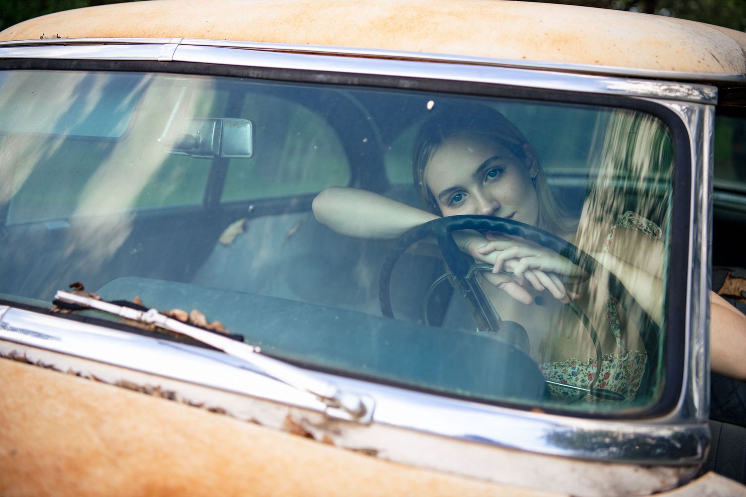 Person gazes dreamily through windshield of vintage car in soft natural light.